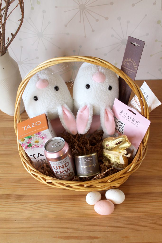 a straw easter basket sits on a table, filled with bunny slippers, tea, a chocolate bunny, lotion, wine, a candle, incense, a face mask, and decorative eggs