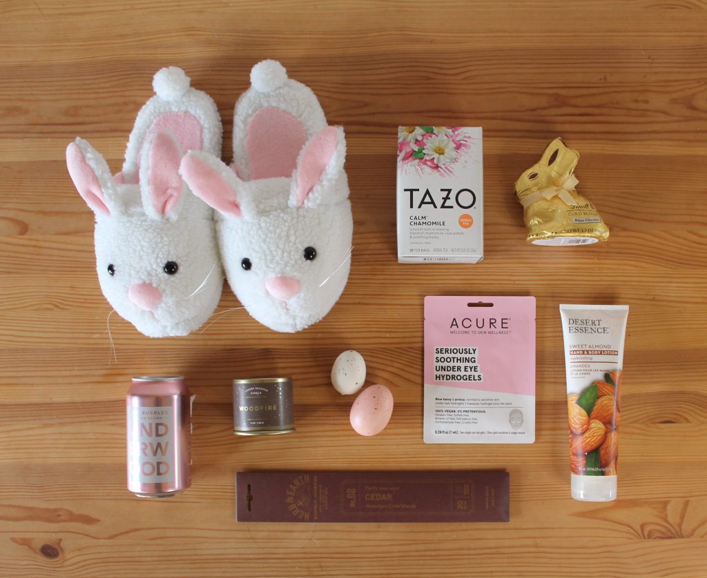 items from an adult easter basket laid out on a wooden table:  bunny slippers, tea, a chocolate bunny, lotion, wine, a candle, incense, a face mask, and decorative eggs