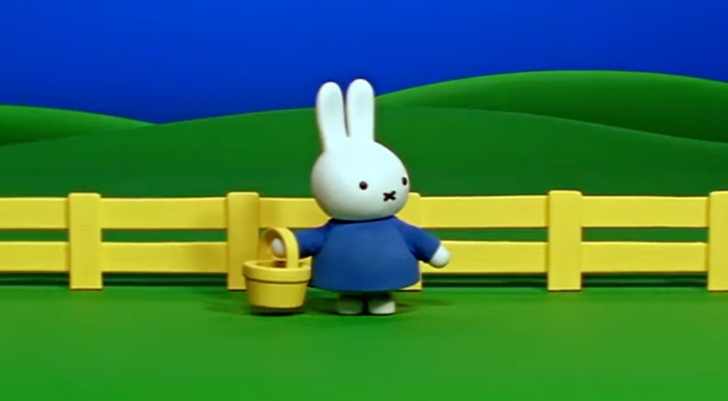 miffy stands in front of a yellow fence holding a basket.  behind her are rolling green hills.