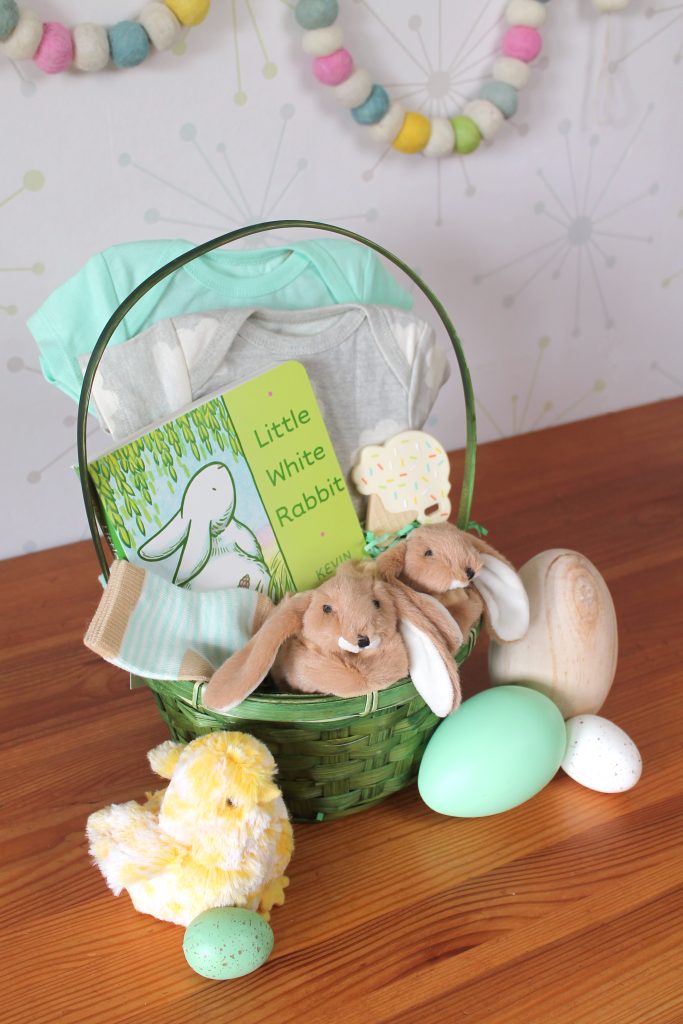 Green Easter basket containing onesies, a book about a bunny, a teether shaped like an ice cream cone, a pair of baby socks, and a pair of brown bunny baby booties.  In front of the basket, there are pastel Easter eggs and a plush baby chick.