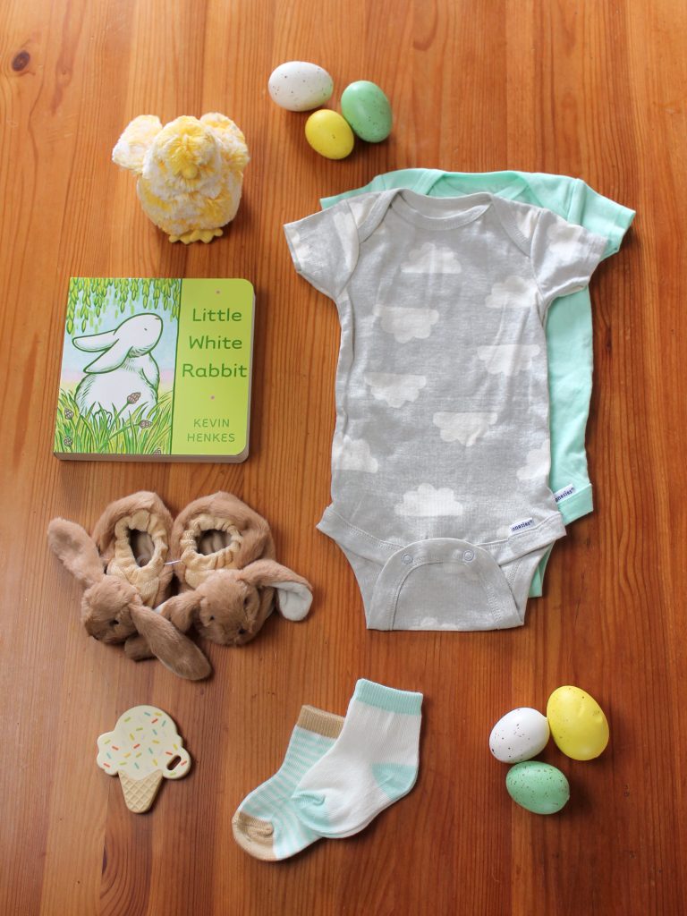 These items are laid out on a wooden table:  two baby onesies, a book about a bunny, a teether shaped like an ice cream cone, a pair of baby socks, and a pair of brown bunny baby booties, pastel Easter eggs and a plush baby chick.