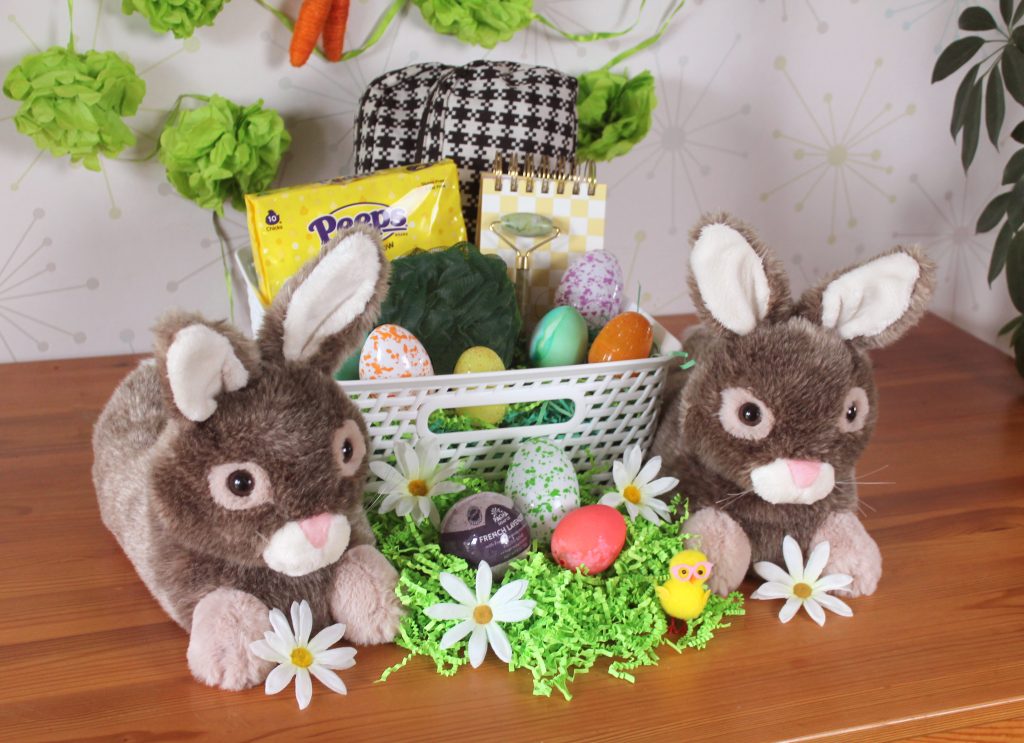 Easter basket for teen girl on top of wooden table.  Basket contains fanny pack, peeps, a notebook, a jade roller, a shower poof, and easter eggs.  A pair of brown bunny slippers is next to the easter basket, and there is a nest made out of easter grass.