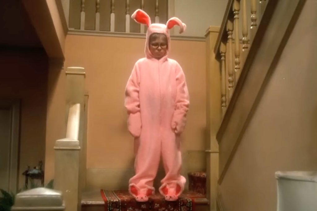 from the movie A Christmas Story, a young boy stands on a staircase wearing a pink bunny costume with pink bunny slippers, and has a very sad expression on his face.
