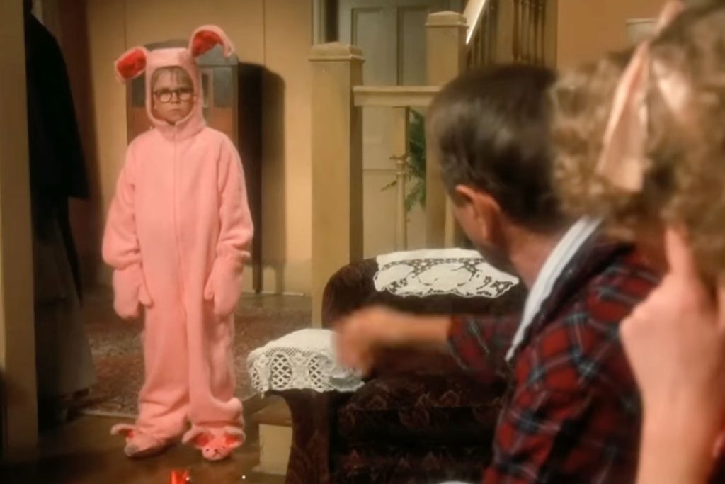 From the movie A Christmas Story, a young boy stands in a living room in front of his parents, wearing a pink bunny suit with bunny slippers and an annoyed expression on his face.