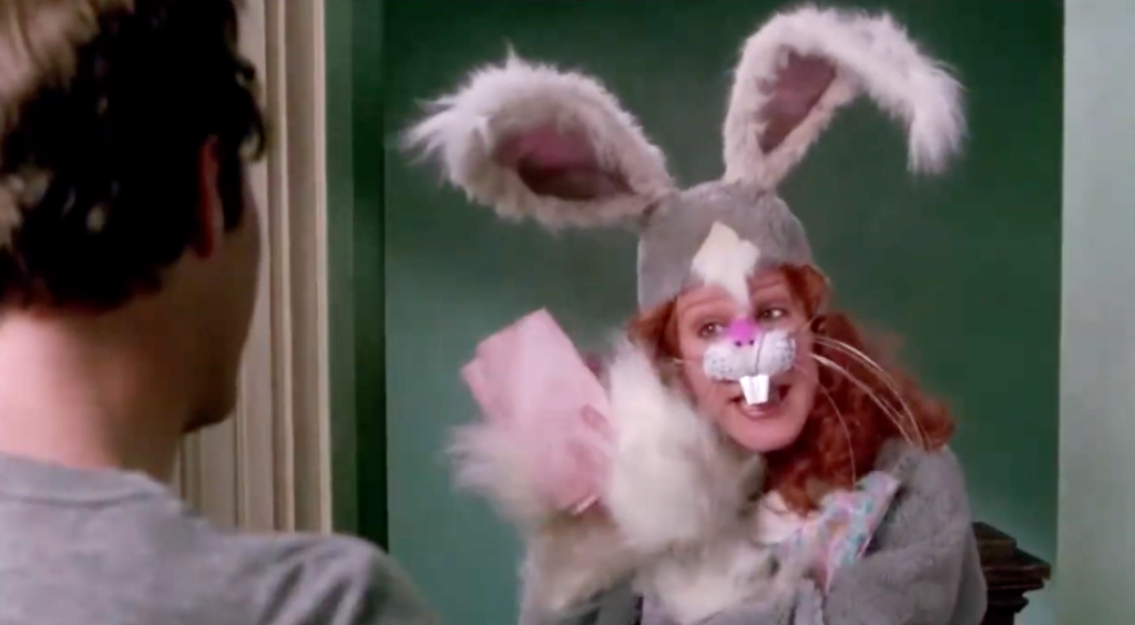 A woman with red hair wears a gray bunny suit and nose with buck teeth, and sings to a man in a doorway.