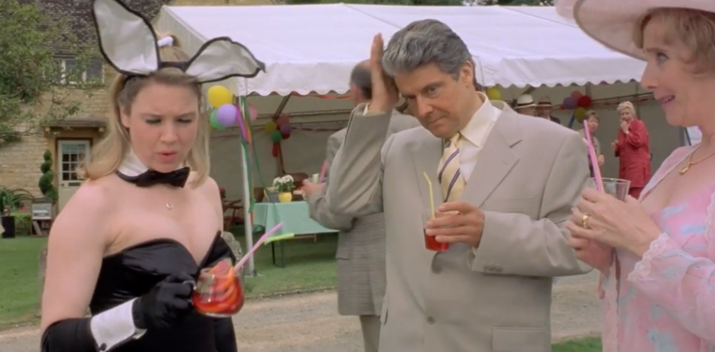 from the movie Bridget Jones Diary, character Bridget stands outdoors at a party holding a cocktail and wearing a playboy bunny outfit. A man in a gray suit and a woman in a pink dress and sun hat stand looking at her.