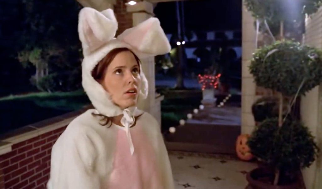 A woman wearing a white and pink bunny costume stands at the door of a house decorated for Halloween.