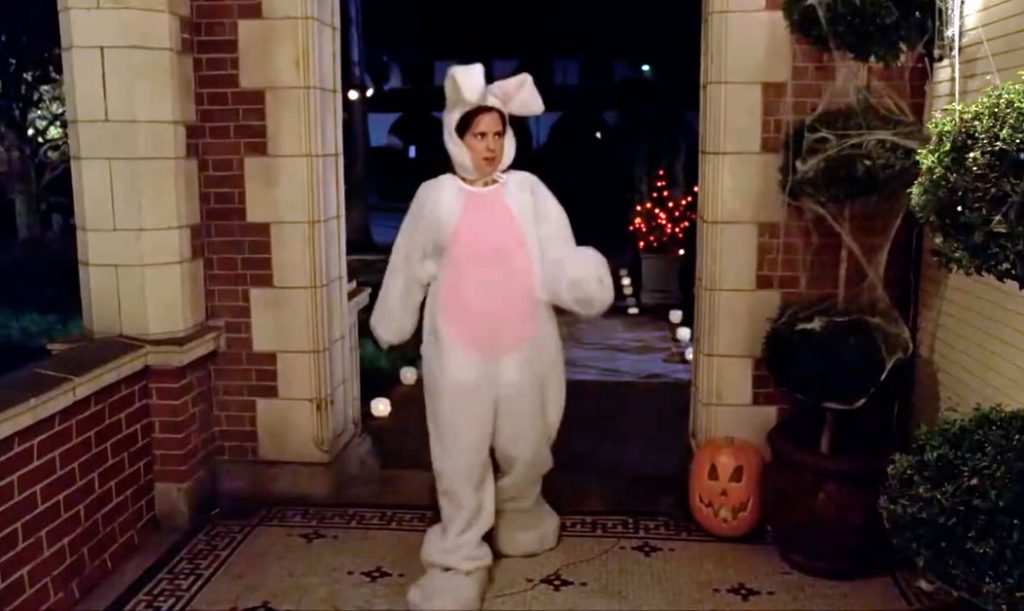 A woman in a full body bunny costume with big furry feet walks into a doorway that is decorated for halloween.