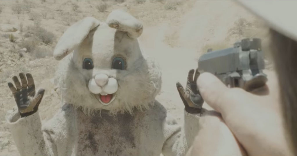 Someone dressed in a dirty Easter Bunny costume holds up their hands while hands point a gun at them.