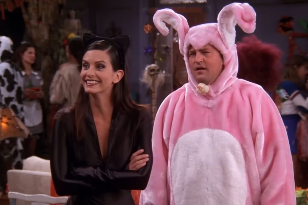 friends tv show characters are at a halloween party in an apartment. monica geller wears a catwoman suit and chandler bing wears a big pink bunny suit with a nose.