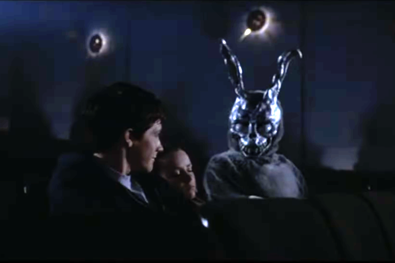 two young people sit in a dark movie theater while a scary looking bunny face looks over at them.
