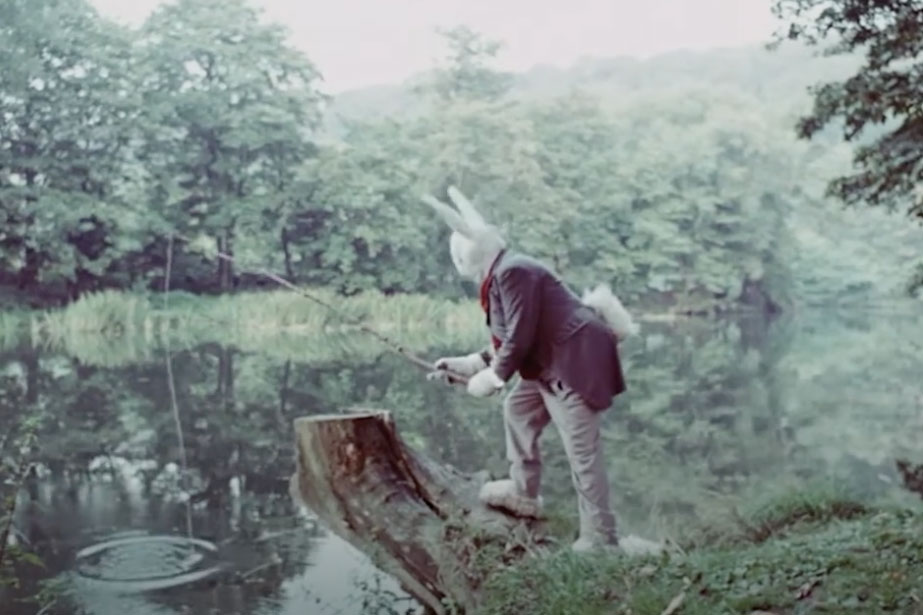 A man in a white bunny hat, gloves and feet wears a suit and goes fishing in a pond.