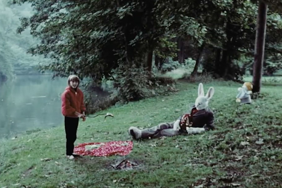 A man in a white bunny head, paws, and feet wears a suit and lays in the grass by a lake. A girl cooks a fish over an open fire.