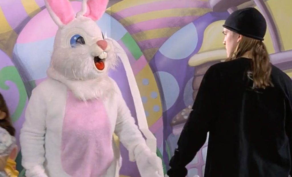 Someone in a white and pink Easter Bunny costume stands in front of a pastel easter egg background, while a man in a black long sleeve t-shirt, black beanie hat and long hair looks at the bunny aggressively.