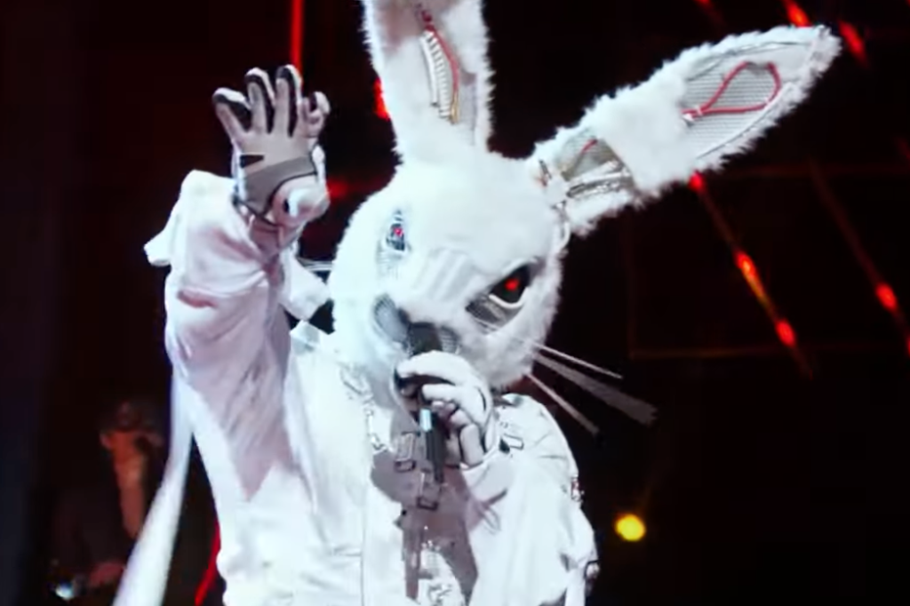 A person wears a bunny costume with a full head covering mask and gloves, and holds a microphone.