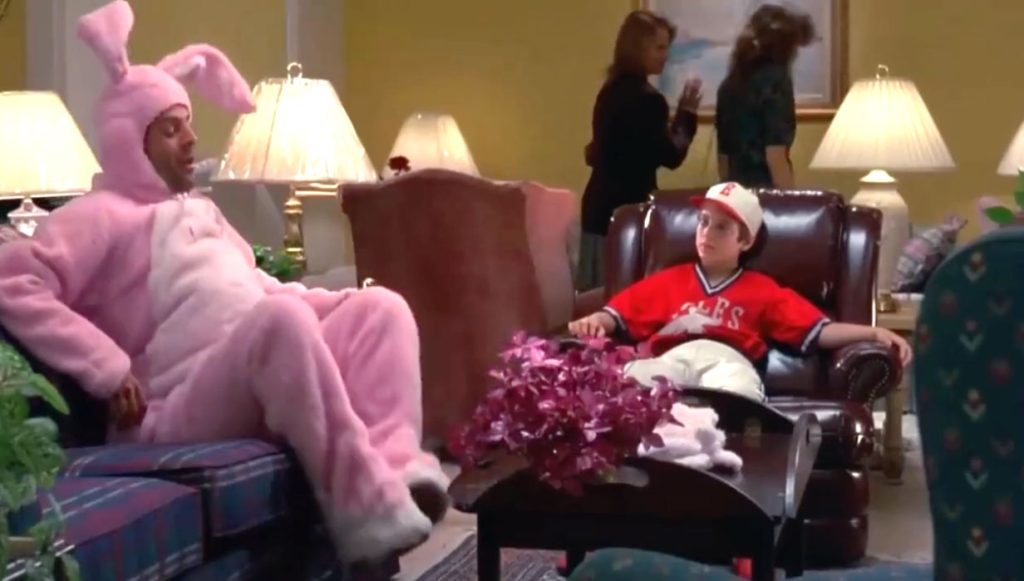A man in a pink bunny suit sits on a plaid couch and a boy in a red baseball uniform sits on a brown leather chair.