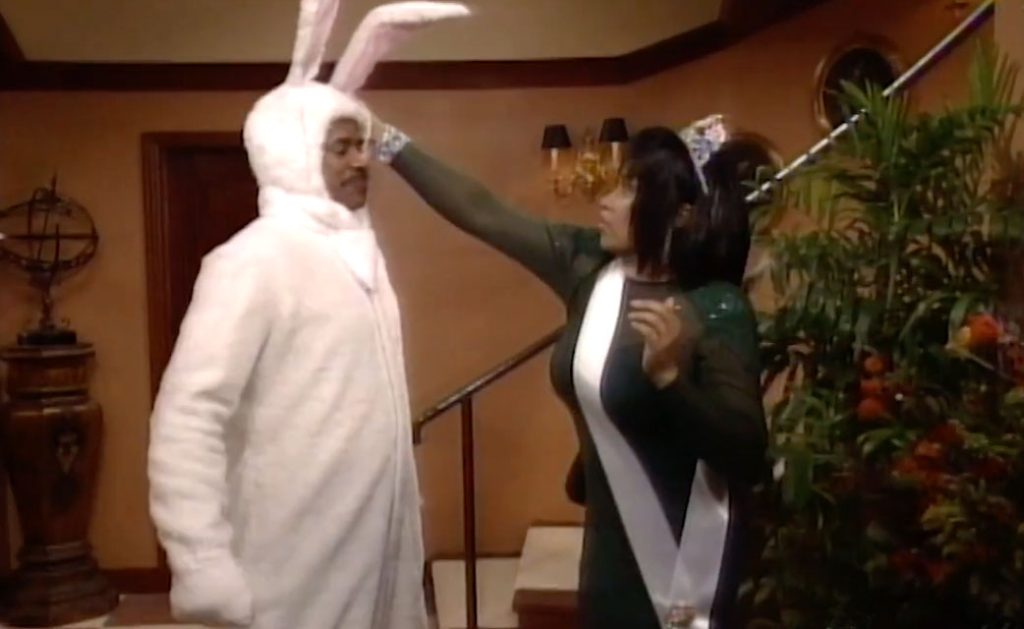 A man in a white bunny suit and a woman in a beauty queen costume stand in a lobby.