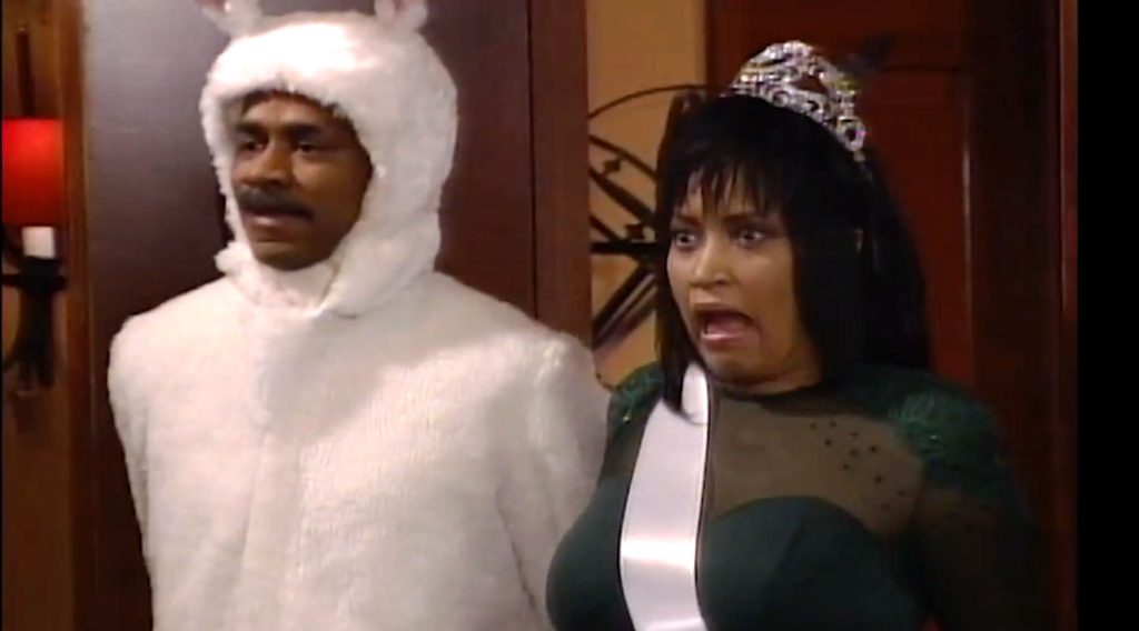A man in a white bunny suit and a woman in a beauty queen costume stand in a room with dark wood paneling and look surprised.